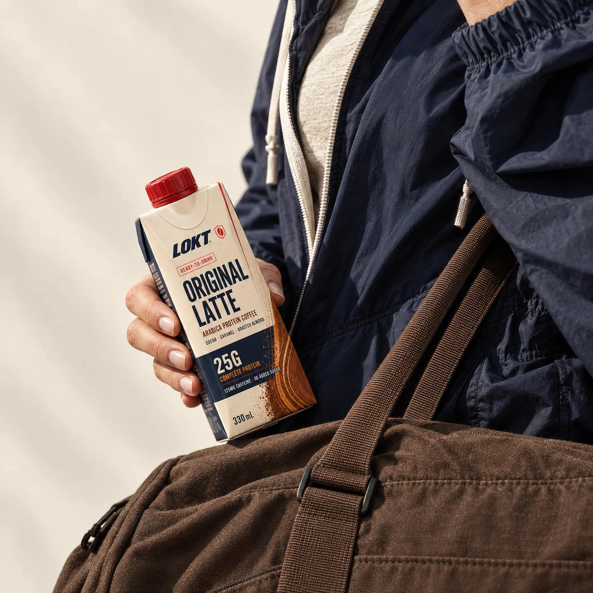 LOKT READY Original Latte held beside a brown gym bag and navy windbreaker