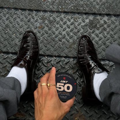 A closed Dark Espresso tin held between grey trousers and polished loafers on worn city steps.