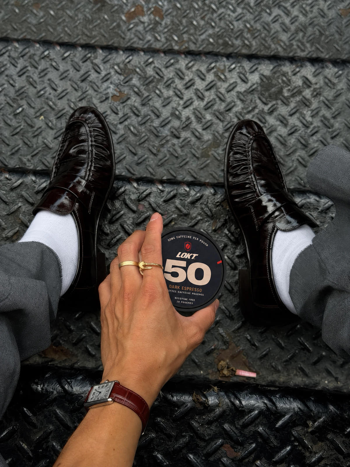 A closed Dark Espresso tin held between grey trousers and polished loafers on worn city steps